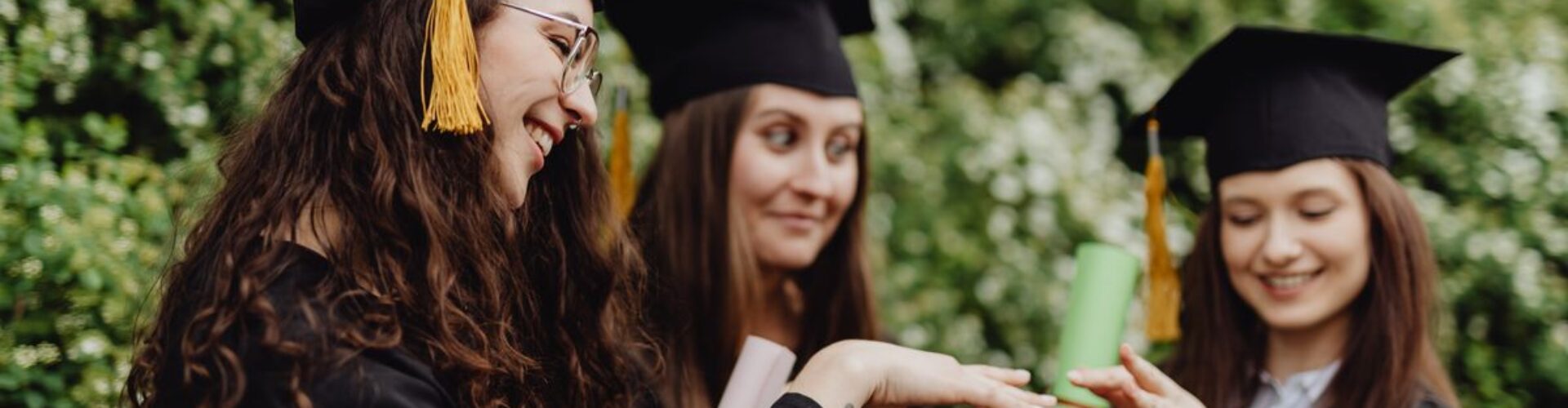 Three young women celebrate graduation outdoors with caps and gowns, showcasing their diplomas and happiness.